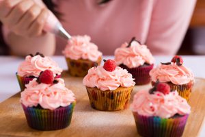 Close shot of many sweet cupcakes on the foreground while a baker decorating the last one