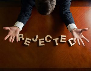 Word Rejected made of wooden block letters and devastated middle aged caucasian man in a black suit sitting at the table, top view composition with dramatic lighting