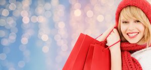 winter holidays, christmas and people concept - smiling young woman in hat and scarf with red shopping bags over lights background